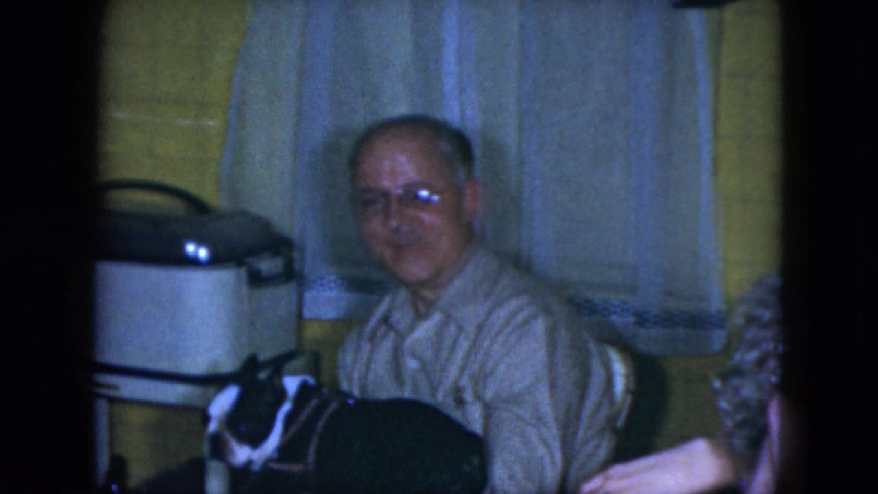 Older man in glasses seated at a kitchen table with a black-and-white dog nearby.