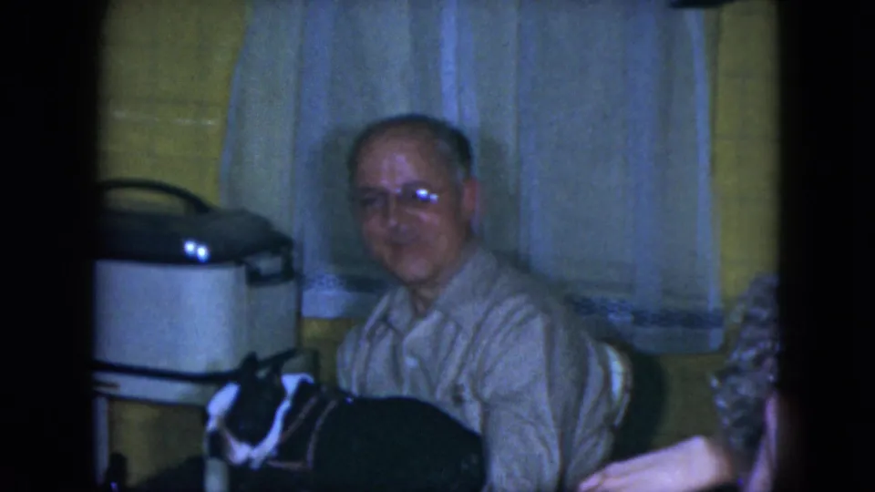Older man in glasses sits in a Cleveland kitchen setting with a Boston Terrier near the table in 1951 home-movie footage.