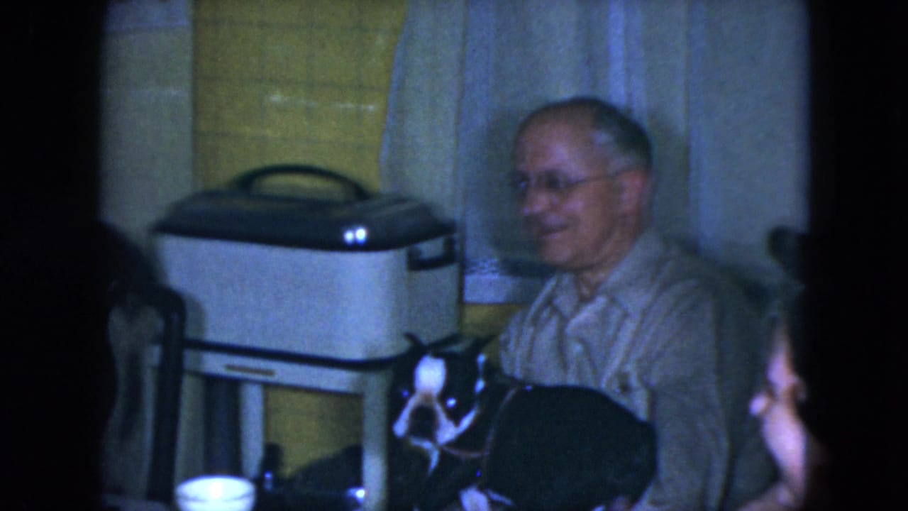 Older man smiling beside a black-and-white dog in a tiled kitchen interior.