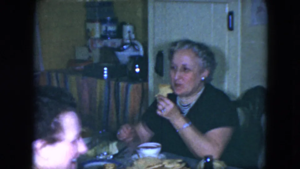 Woman holds food at a Cleveland kitchen table while other adults remain partly visible in 1951 home-movie footage.