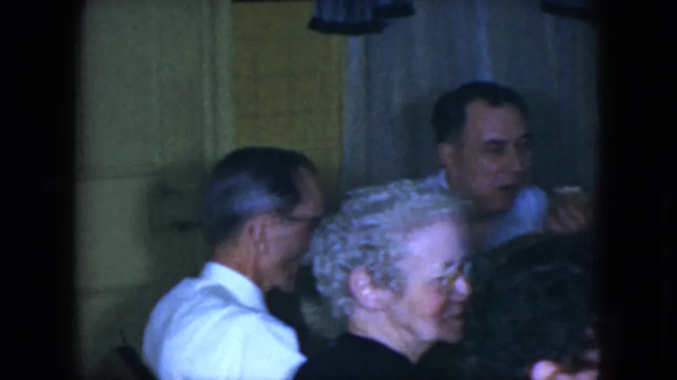 1951 Cleveland home-movie frame showing adults at a private dining table, with cups, food, curtains, and soft indoor lighting visible.