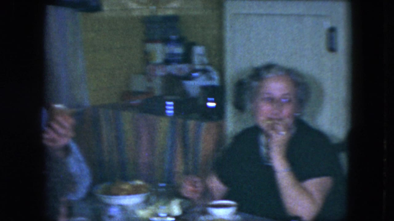 An elderly woman sits at a table with dishes and a cup nearby.