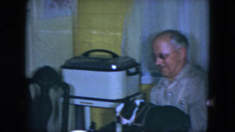An older man sits beside a dining table in the 1951 Cleveland kitchen, near a white domestic appliance and curtained window.
