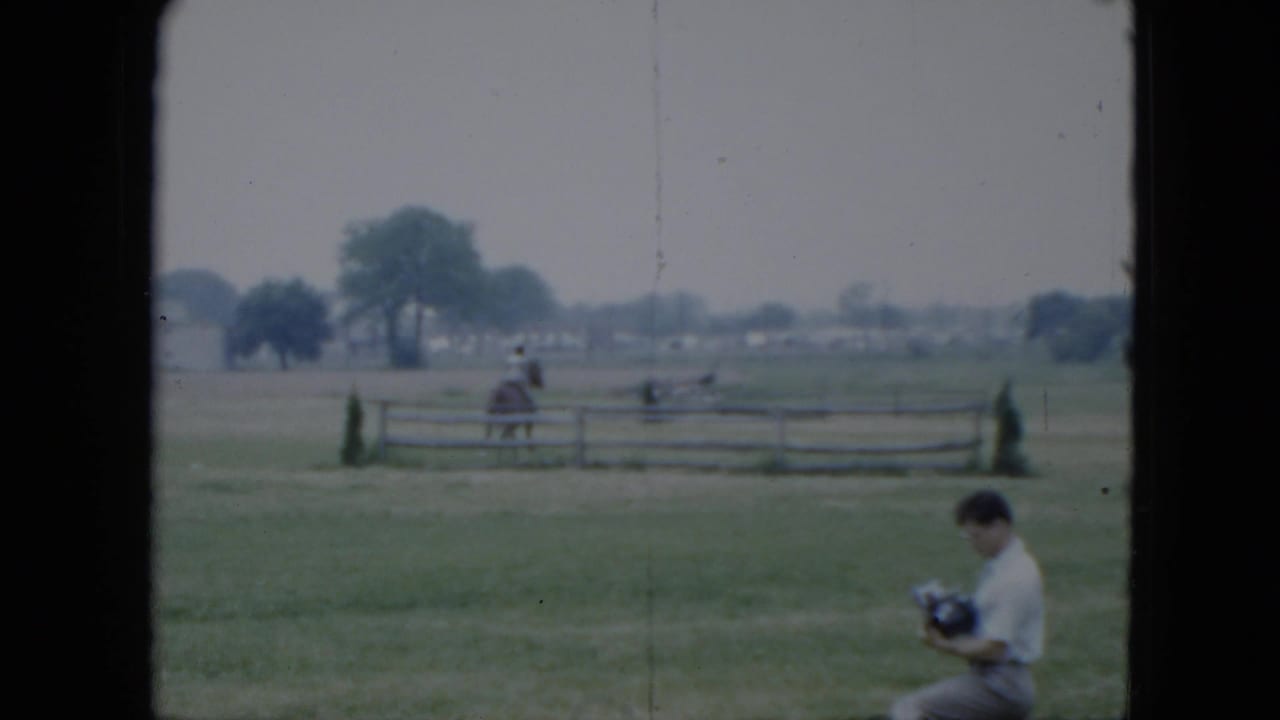 Seated man in a field as a horseback rider passes behind a wooden fence.