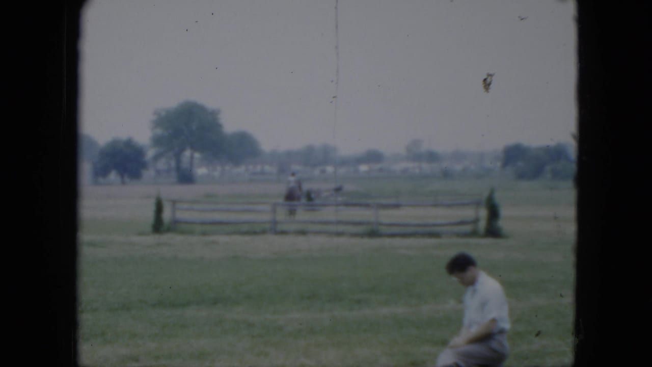 Man sits at the edge of a grassy field with a rider and fence in the background.