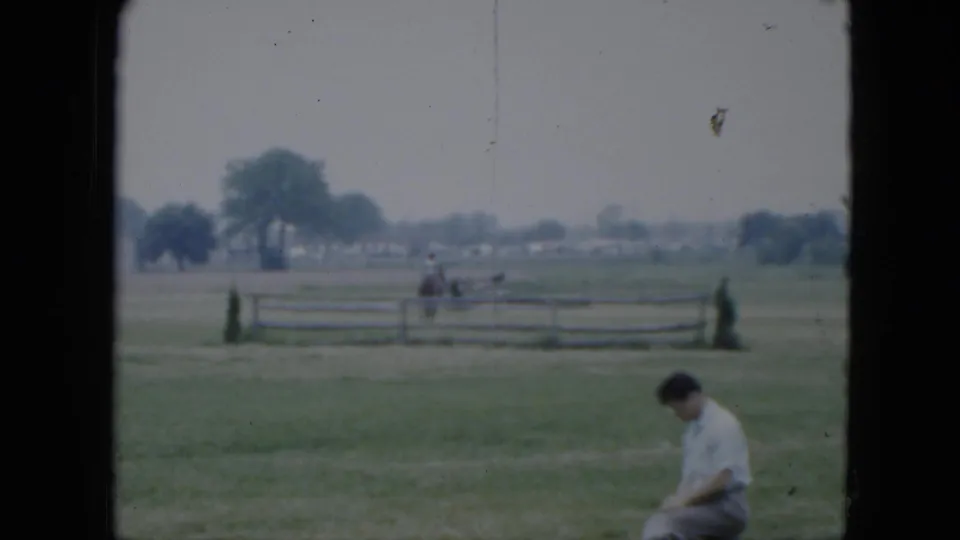1951 Cleveland-area home-movie frame with a seated man at right and a horseback rider crossing the field behind a fence.