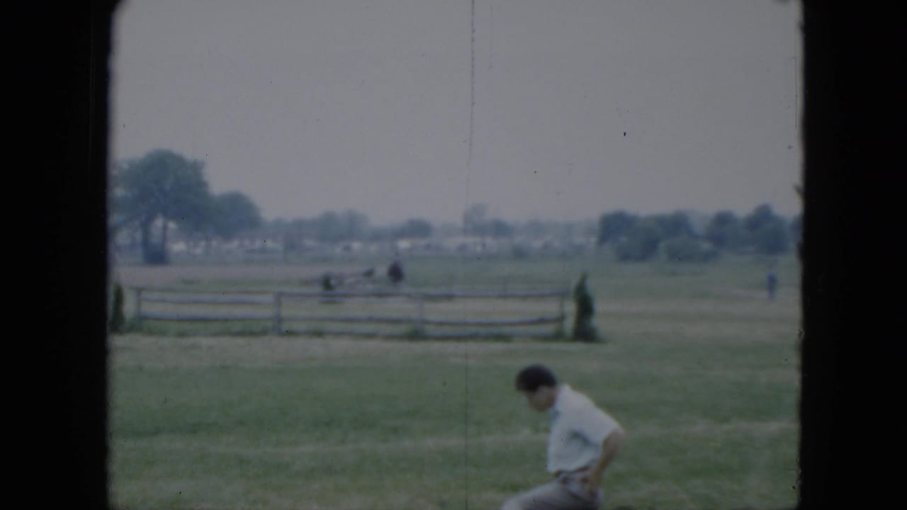 Grassy fairground with a seated person near the foreground and rail fencing in the distance.