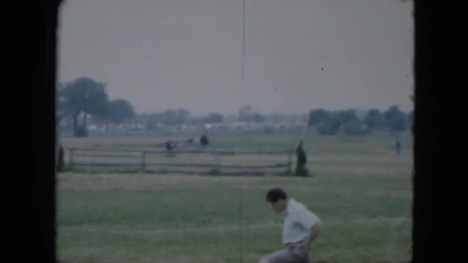 1951 Cleveland home-movie frame showing a person seated on grass near a fairground field, with fencing and trees beyond.