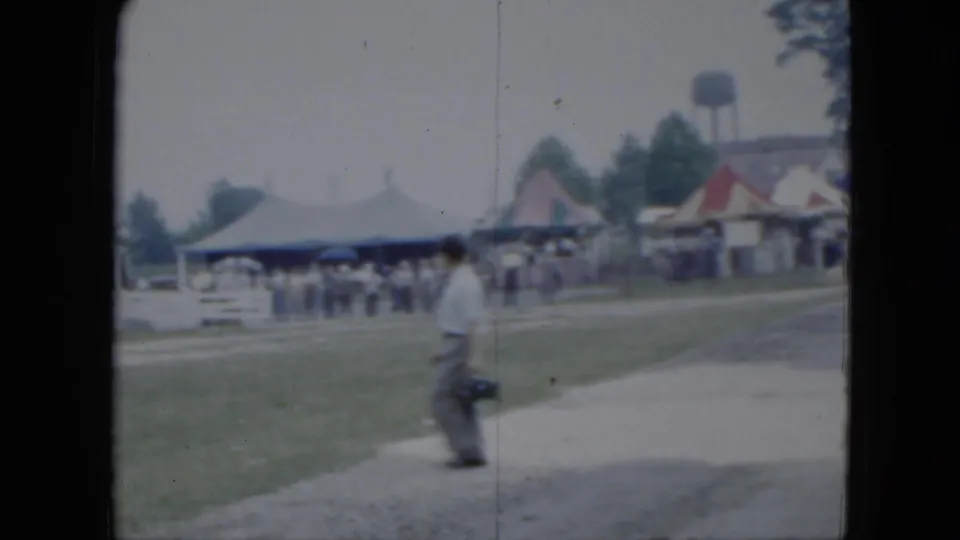 1951 Cleveland fairground frame with a man carrying a camera near tents, spectators, a path, and a distant water tower.