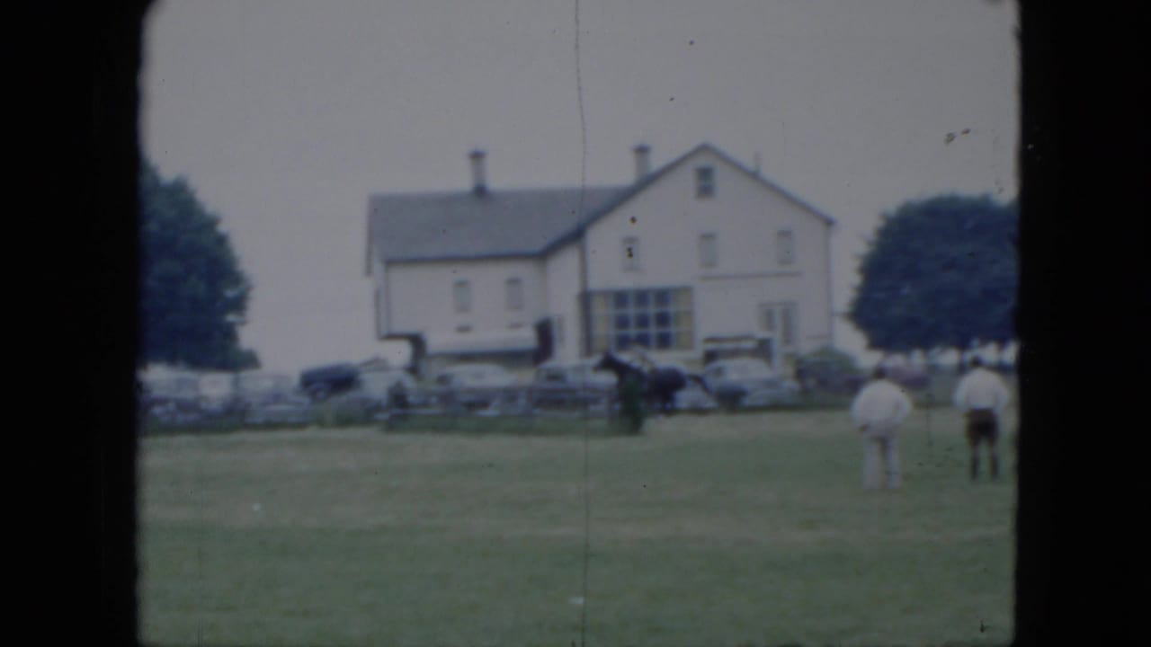 Dark horse and rider crossing a grassy field near spectators, parked cars, and a white building.