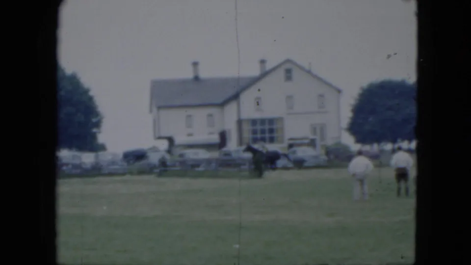 1951 Cleveland horse-show frame with a dark horse and rider on grass, spectators in light clothing, period cars, and a white building behind them.