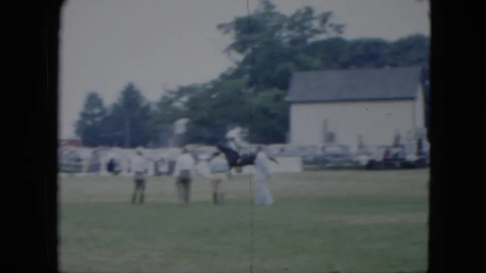 1951 Cleveland horse-show frame with the rider and dark horse near the foreground, several spectators standing nearby, and a white building beyond the field.