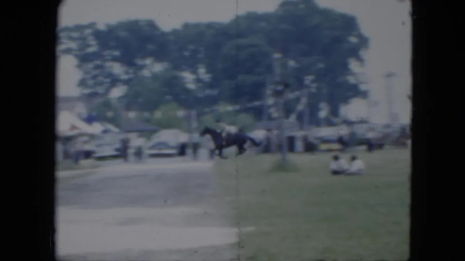 1951 Cleveland home-movie frame showing a mounted rider moving across grass near spectators, parked cars, and trees.