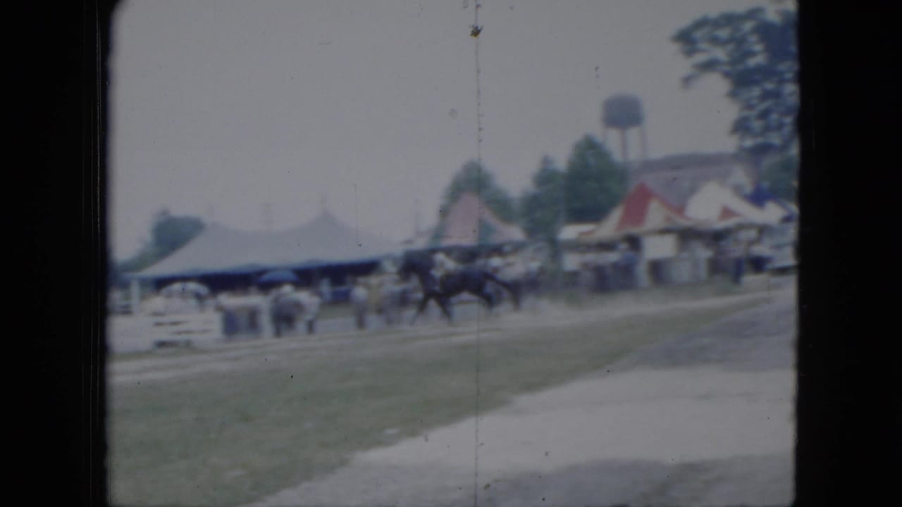 1951 Cleveland fairground view with a rider on horseback passing tents or pavilion roofs as spectators stand nearby.