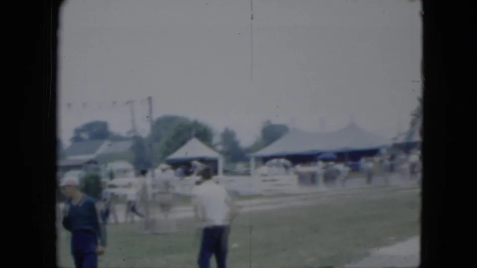 1951 Cleveland home-movie frame showing spectators near a riding area with horses, pavilion structures, and cars behind them.