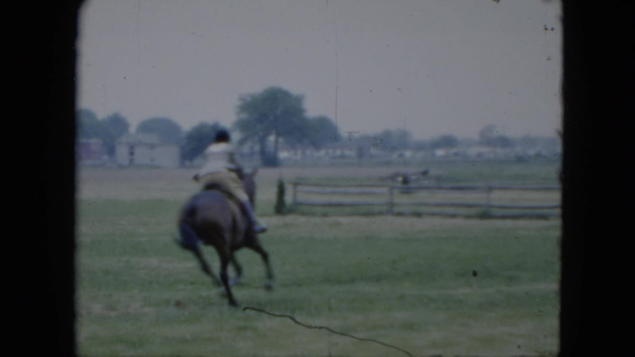 1951 Cleveland home-movie frame showing a rider on a dark horse moving across an open grassy field with a rail fence and distant trees.