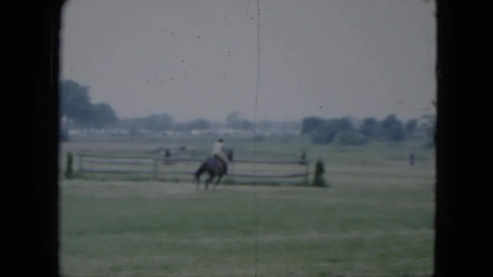 A soft 8mm frame shows the rider and horse near the rail fence, with open grass and distant tree shapes in the background.