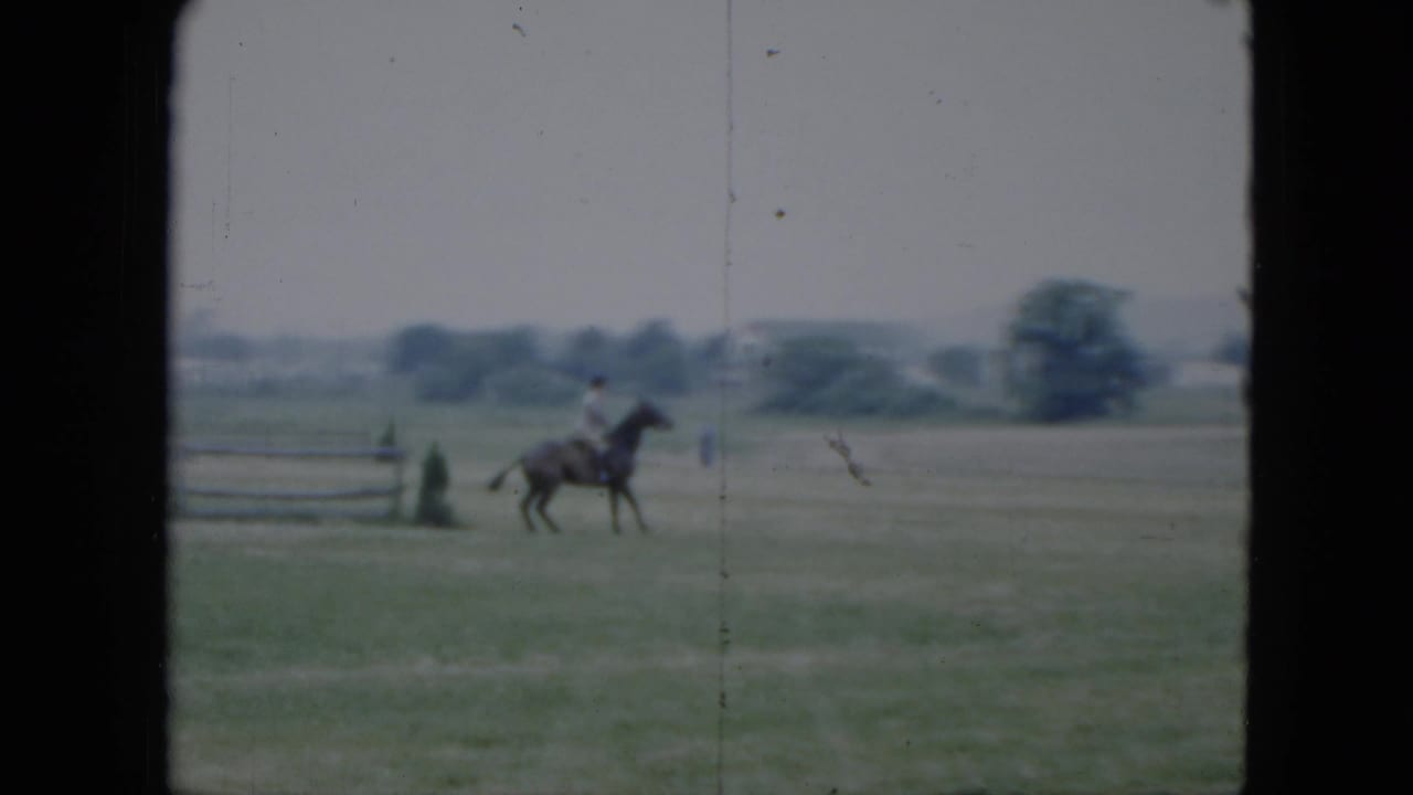 1951 Cleveland home-movie frame showing a distant rider on horseback in an open field with fence rails and houses beyond.