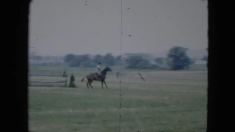 1951 Cleveland home-movie frame showing a distant rider on horseback in an open field with fence rails and houses beyond.