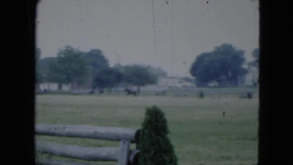 1951 Cleveland 8mm frame of a distant rider and horse moving across a grassy field with trees, houses, fence, and shrub in view.