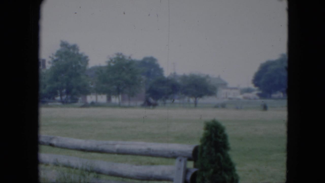Grassy field behind a split-rail fence with trees and distant buildings.