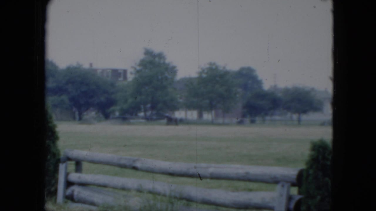 1951 Cleveland home-movie frame showing open grass, a split-rail fence, trees, and faint buildings in the distance.