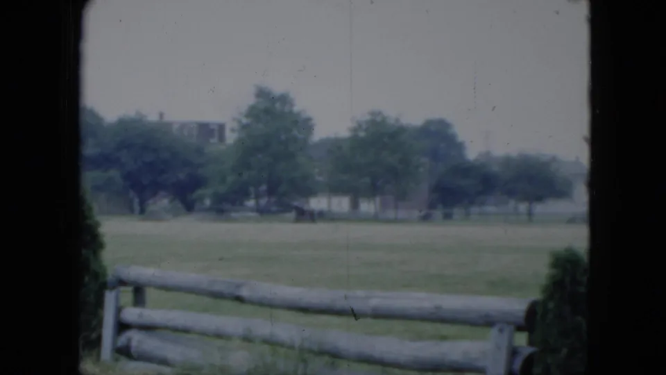 1951 Cleveland home-movie frame showing open grass, a split-rail fence, trees, and faint buildings in the distance.