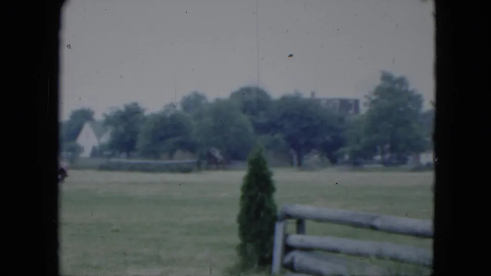 1951 Cleveland home-movie frame of a distant rider on horseback near trees, viewed across grass and foreground fence rails.