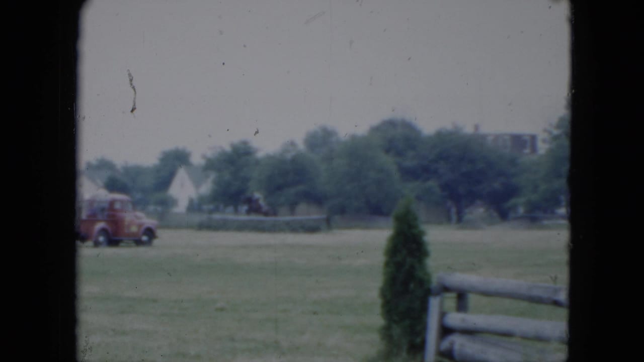 Red truck parked beyond a grassy field with trees, houses, fence, and film gate edges visible.