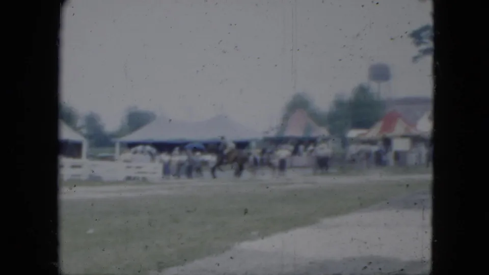 1951 Cleveland-area 8mm frame showing people and horses near a field edge, with low buildings, houses, and a water tower in the distance.