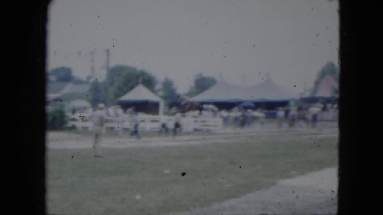 Grassy equestrian field with white fencing, tents, and distant spectators.