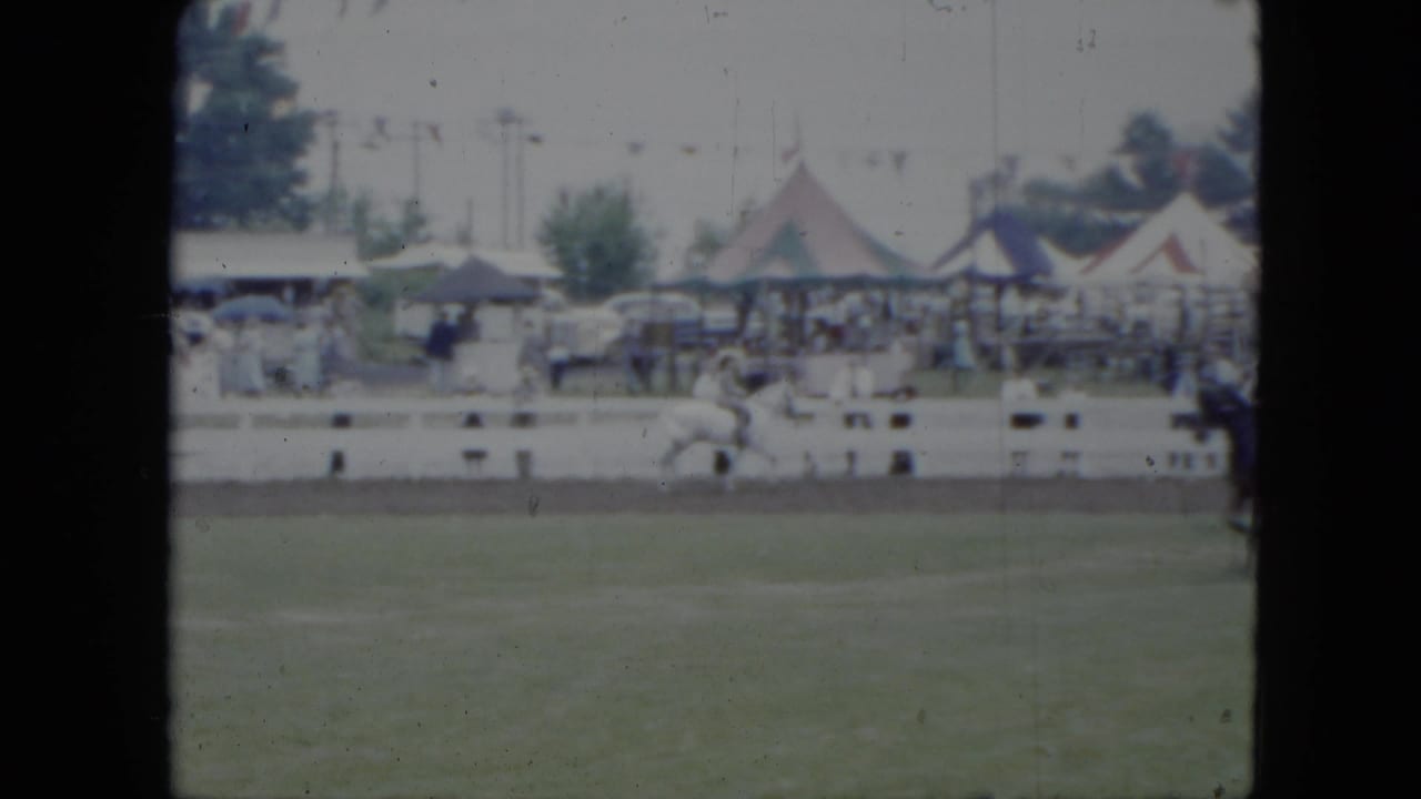 1951 Cleveland fairground footage showing a rider on a white horse in a grassy arena, with striped tents, spectators, and parked cars beyond the rail.