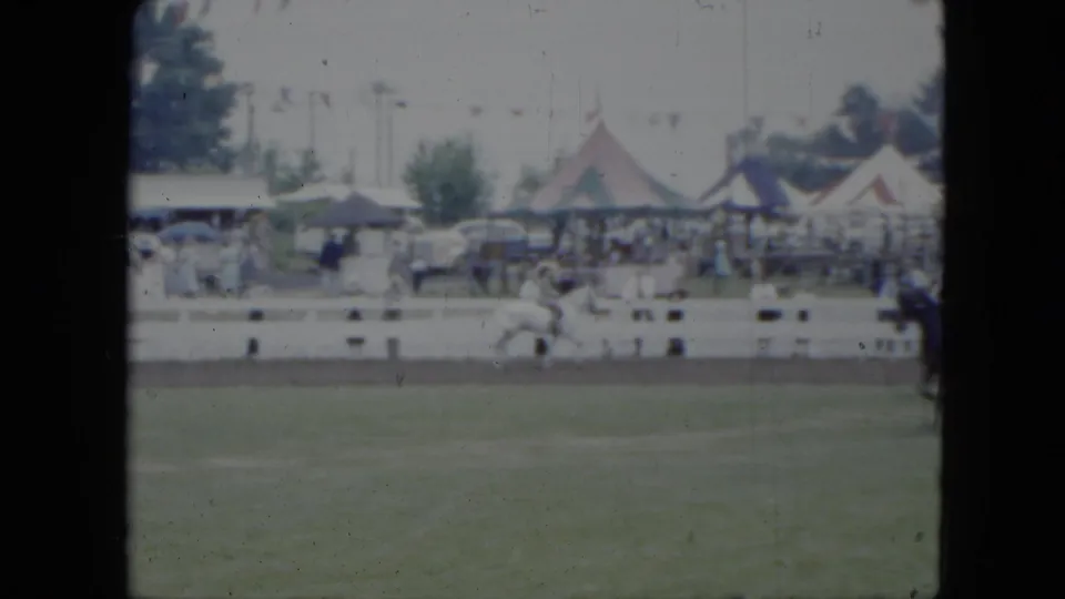 1951 Cleveland fairground footage showing a rider on a white horse in a grassy arena, with striped tents, spectators, and parked cars beyond the rail.