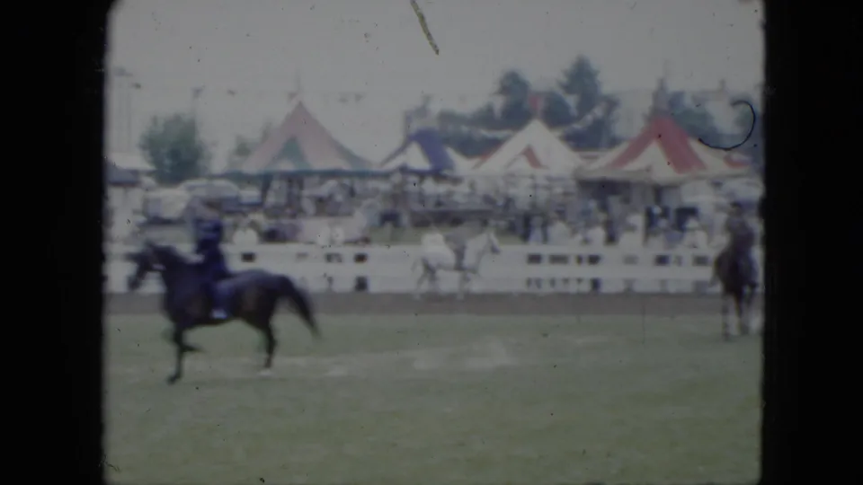 A dark horse and rider move across the Cleveland horse-show ring, with a white horse, tents, spectators, and fairground fencing in the background.