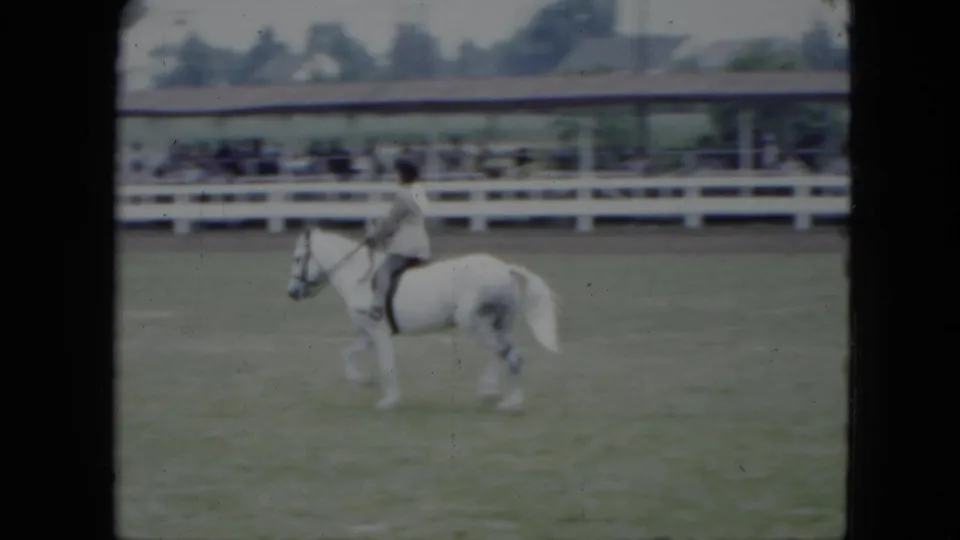 A rider on a white horse crosses the grassy Cleveland fairground arena, with rail fencing and a covered viewing structure in the background.