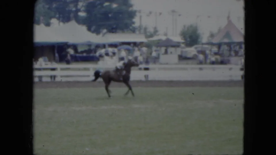 The dark horse and rider continue across the 1951 Cleveland show field, backed by white fencing, tents, trees, and a crowd area.