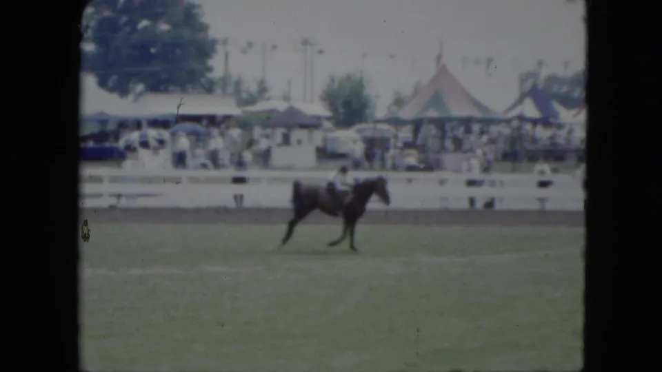 In 1951 Cleveland home-movie footage, a light-clothed rider guides a dark horse through a grassy ring bordered by white fencing and tents.