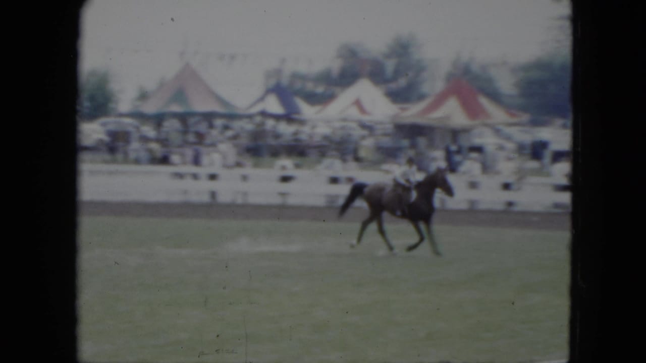 The rider continues around the 1951 Cleveland ring as spectators stand beyond white fencing and colorful tent tops appear in the background.