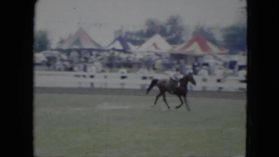 The rider continues around the 1951 Cleveland ring as spectators stand beyond white fencing and colorful tent tops appear in the background.