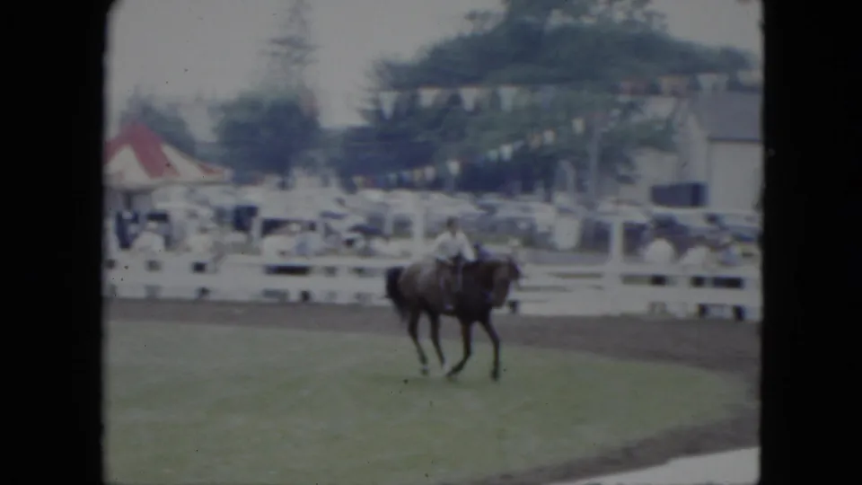 A dark horse and rider angle around the ring in Cleveland, with white fencing, a tent, parked cars, bunting, and trees behind them.