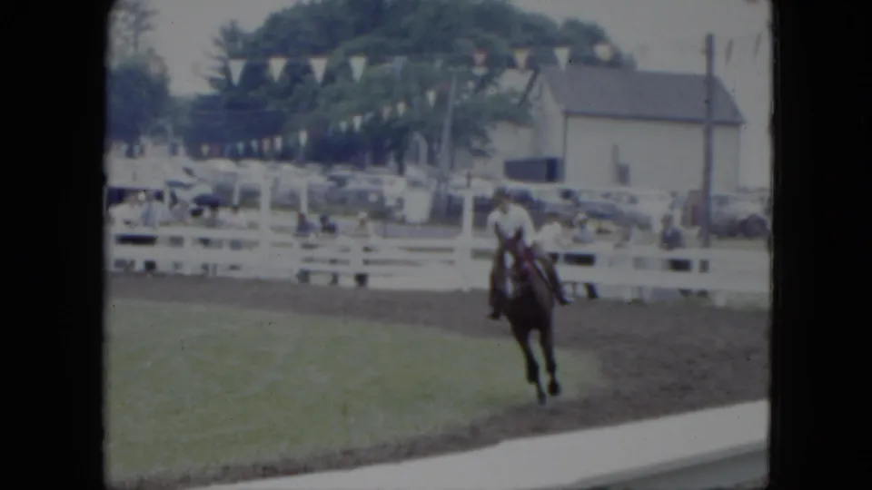 The horse and rider approach the camera side of the 1951 Cleveland ring, with spectators, pennant bunting, white fencing, and nearby buildings visible.