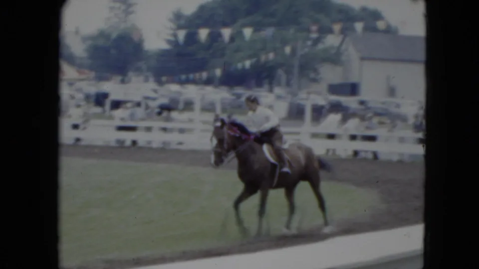 In 1951 Cleveland home-movie footage, a rider in a light shirt guides a brown horse along a grassy equestrian ring.