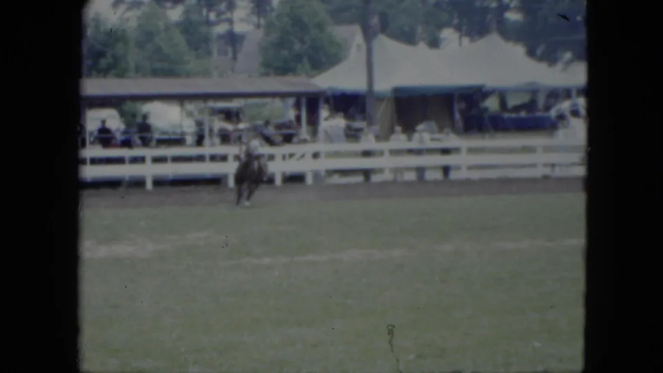 The rider guides the horse across the Cleveland arena while distant spectators sit or stand behind the white rail fence.