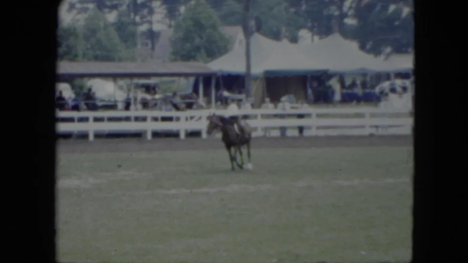 1951 Cleveland home-movie frame showing a rider on a dark horse in a grass ring while spectators watch from behind a white fence.