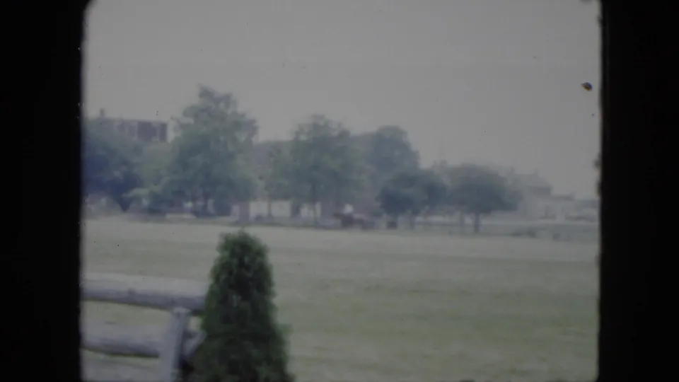 Home-movie view across a Cleveland grassy field, framed by a fence rail, shrub, trees, and houses in the distance.