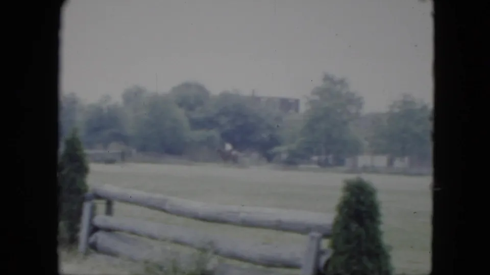 1951 Cleveland 8mm frame of an open field and rail fence, with shrubs, trees, and a distant figure that may be a rider.