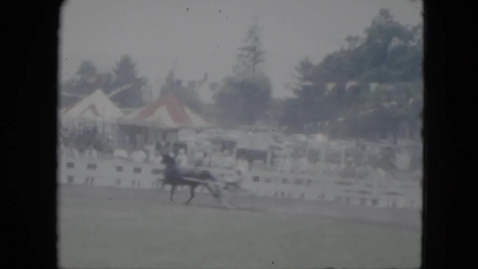 1951 Cleveland home-movie frame of a dark horse pulling a sulky along a grassy track with tents and spectators behind a white fence.