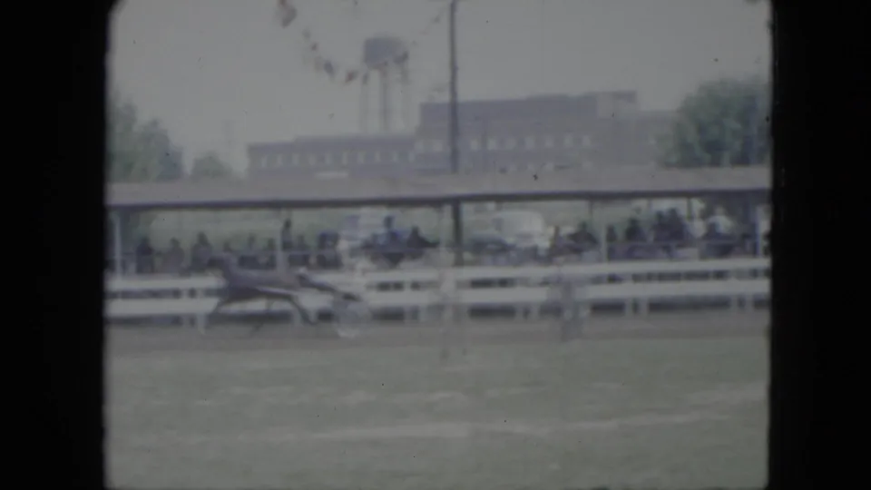 1951 Cleveland home-movie frame of a horse and sulky moving past a fenced spectator area with bunting and a distant brick building.
