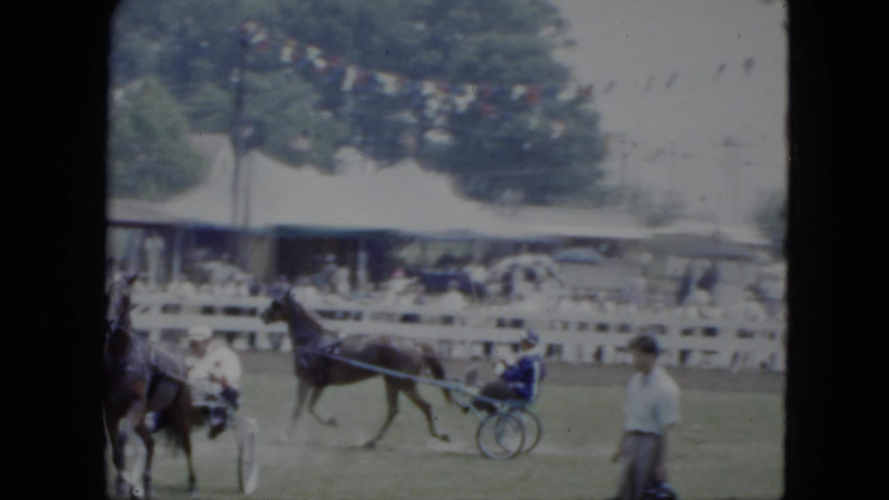 1951 Cleveland fairground view of a harness horse and sulky driver passing a white fence, with spectators, tent shade, trees, and a man near the track.