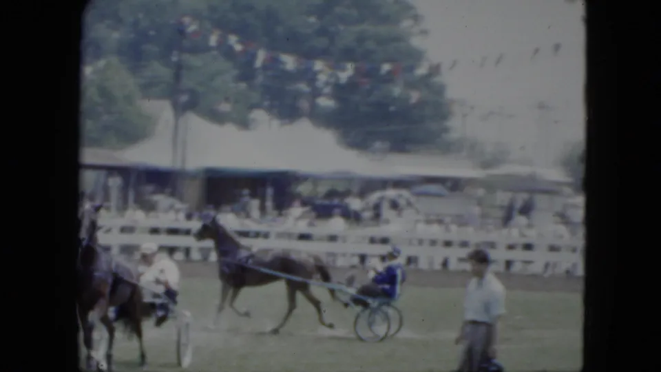 1951 Cleveland fairground view of a harness horse and sulky driver passing a white fence, with spectators, tent shade, trees, and a man near the track.